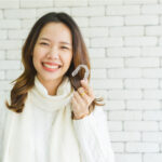 A woman in a white turtleneck smiles while holding her Invisalign clear aligners