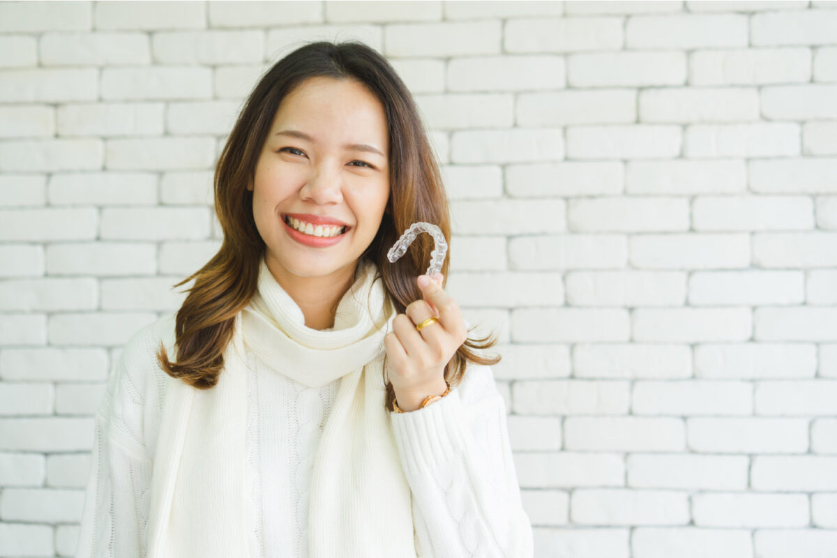 A woman in a white turtleneck smiles while holding her Invisalign clear aligners