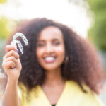 Curly-haired woman in a yellow blouse smiles and holds up her Invisalign aligners outside