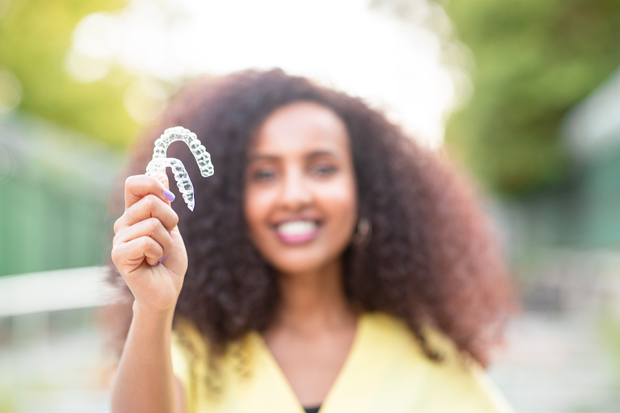 Curly-haired woman in a yellow blouse smiles and holds up her Invisalign aligners outside