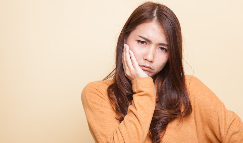 Brunette Asian woman in a golden sweater cringes in pain and holds her cheek due to teeth sensitivity