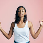 Beautiful Brown woman in a white tanktop stands meditating with her index fingers touching her thumbs against a pink wall