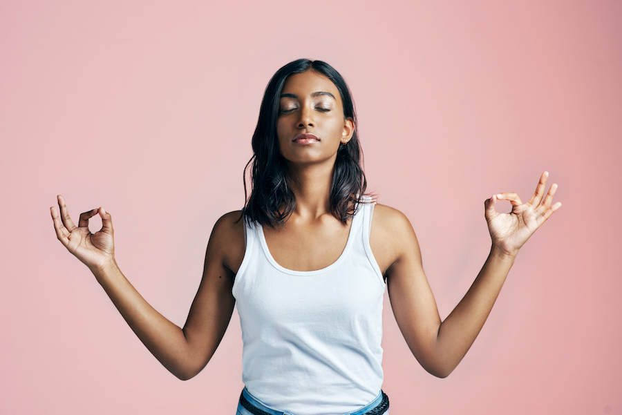 Beautiful Brown woman in a white tanktop stands meditating with her index fingers touching her thumbs against a pink wall