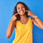Brown woman in a yellow tank top points to her smile after teeth whitening