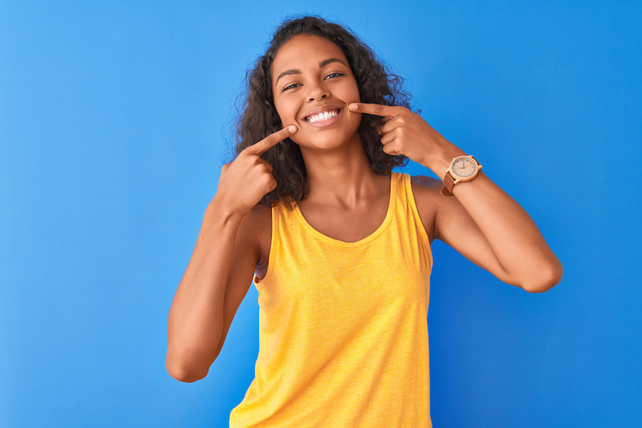 Brown woman in a yellow tank top points to her smile after teeth whitening