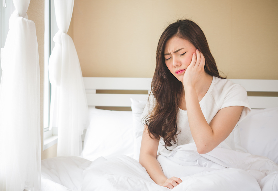 Asian woman cringing in pain and holding her cheek as she sits up in herbed