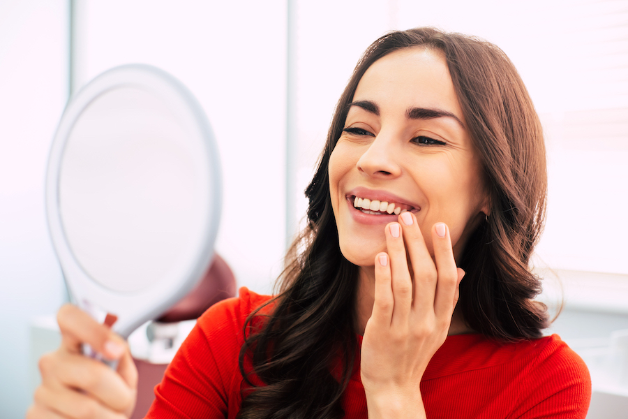 Brunette woman smiles at herself in a handheld mirror after teeth whitening while wearing a red blouse